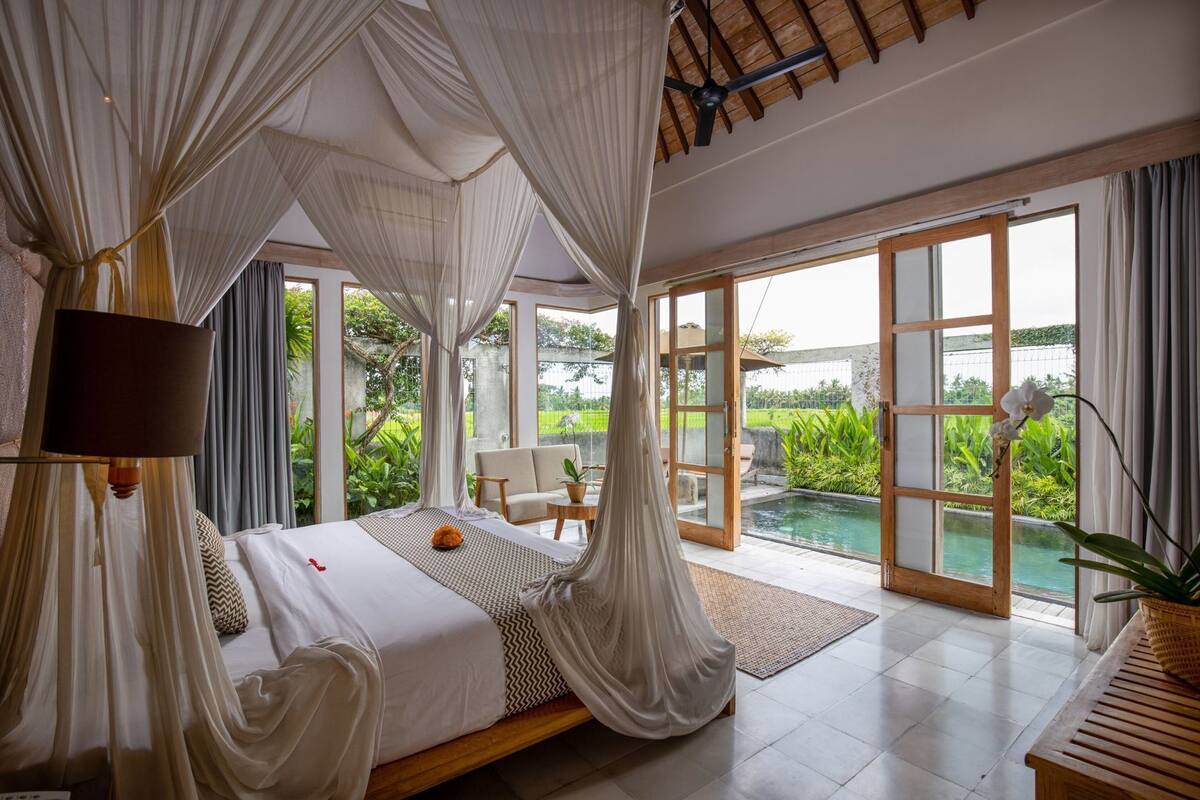Quarto com cama, cortinas, TV e vista para a piscina e jardim.