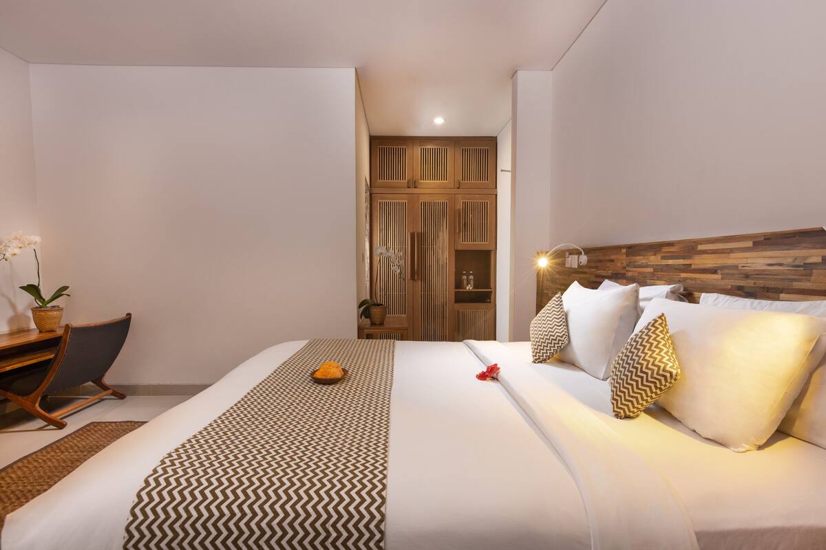 Quarto de hotel com cama, escrivaninha, cadeira, planta em vaso e armário.