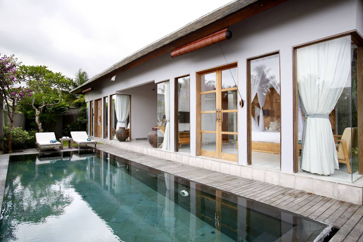 Uma área de piscina moderna com piscina transparente, deck de madeira e duas espreguiçadeiras.