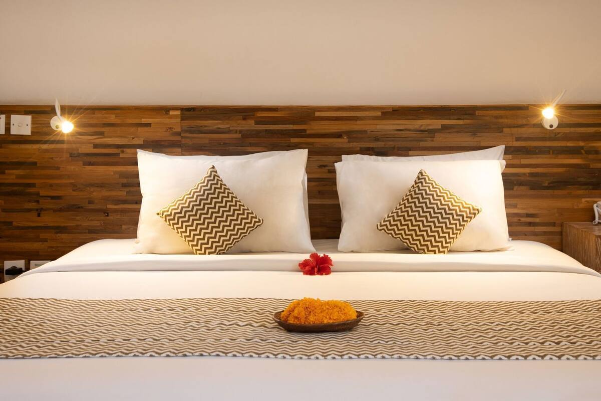 Quarto de hotel com cabeceira de madeira, duas almofadas com padrão geométrico em chevron, colcha branca com textura e uma mesinha com um telefone e uma tigela de frutas.