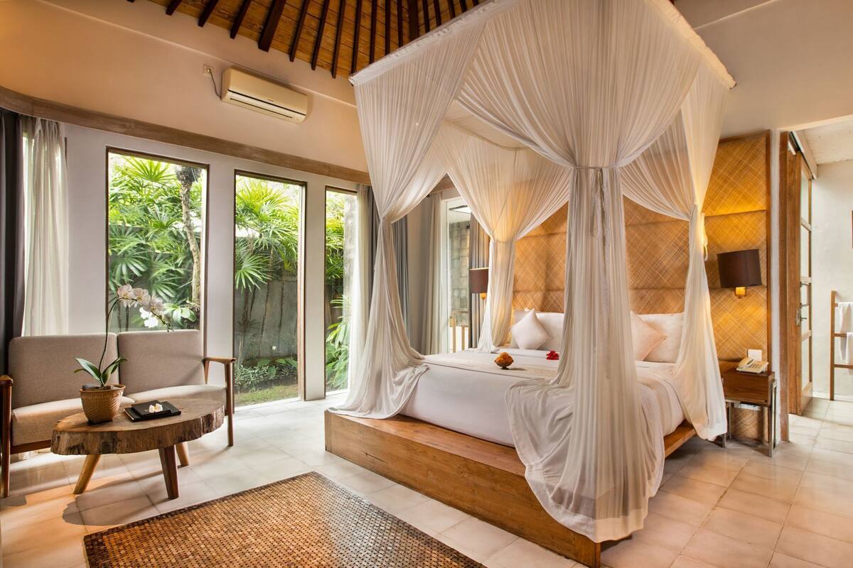 Quarto com cama de dossel, grandes janelas, mesa de cabeceira e vista para o verde.