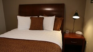 A neatly made bed with a patterned bedspread, a wooden headboard, and a bedside table with a lamp and a clock. - Affordable Suites Kannapolis (Kannapolis)