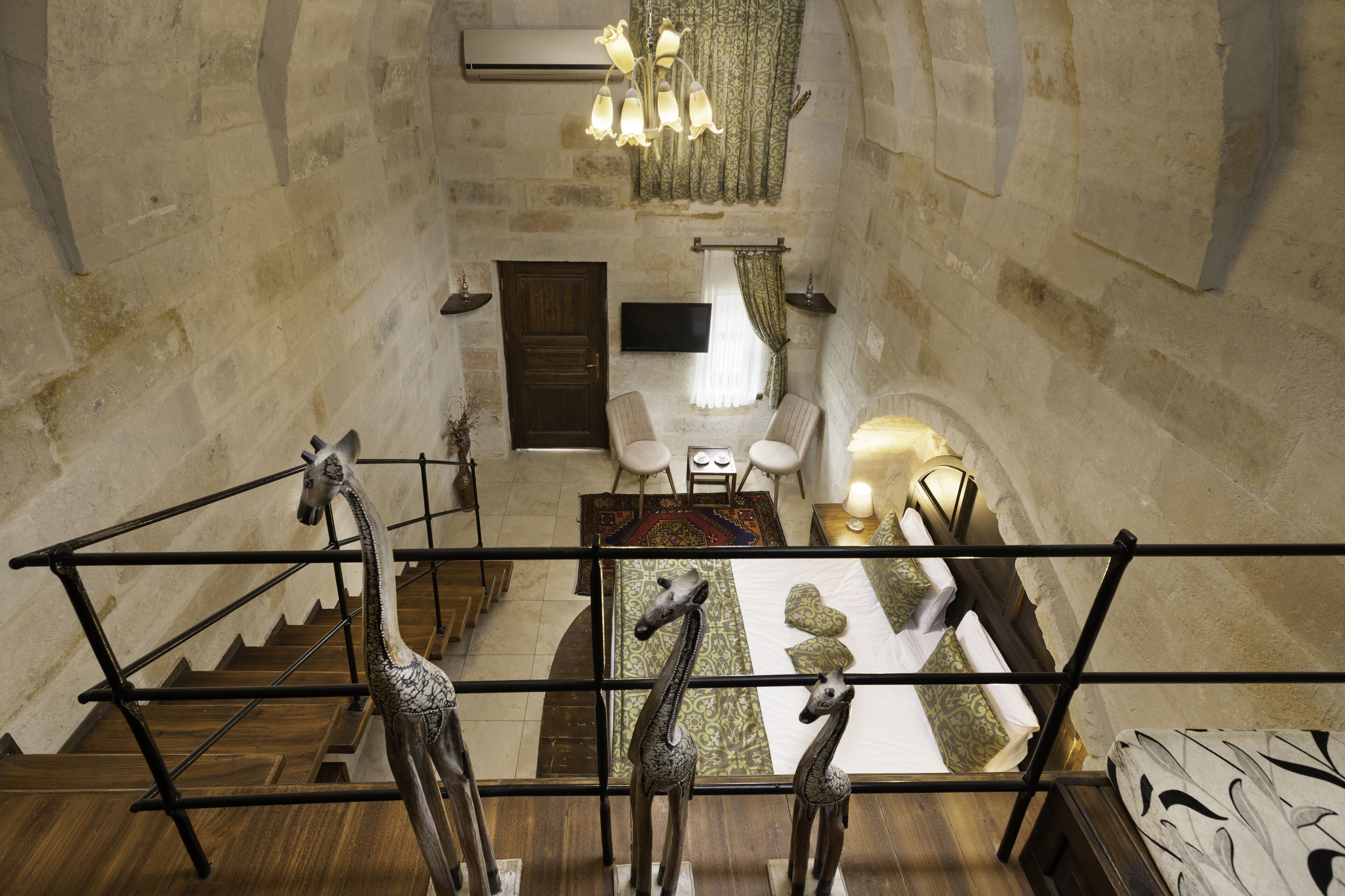 A staircase with wooden railings and a giraffe sculpture at the bottom. There is a bed with patterned bedding and a bedside table with a lamp. A television is mounted on the wall above a window with curtains.