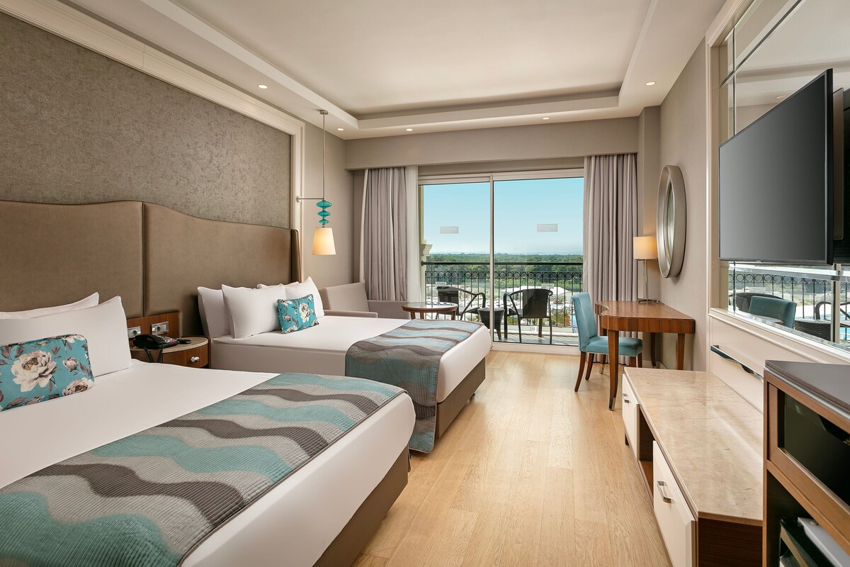 A hotel room with two beds, a desk, a TV, and a balcony with a view.