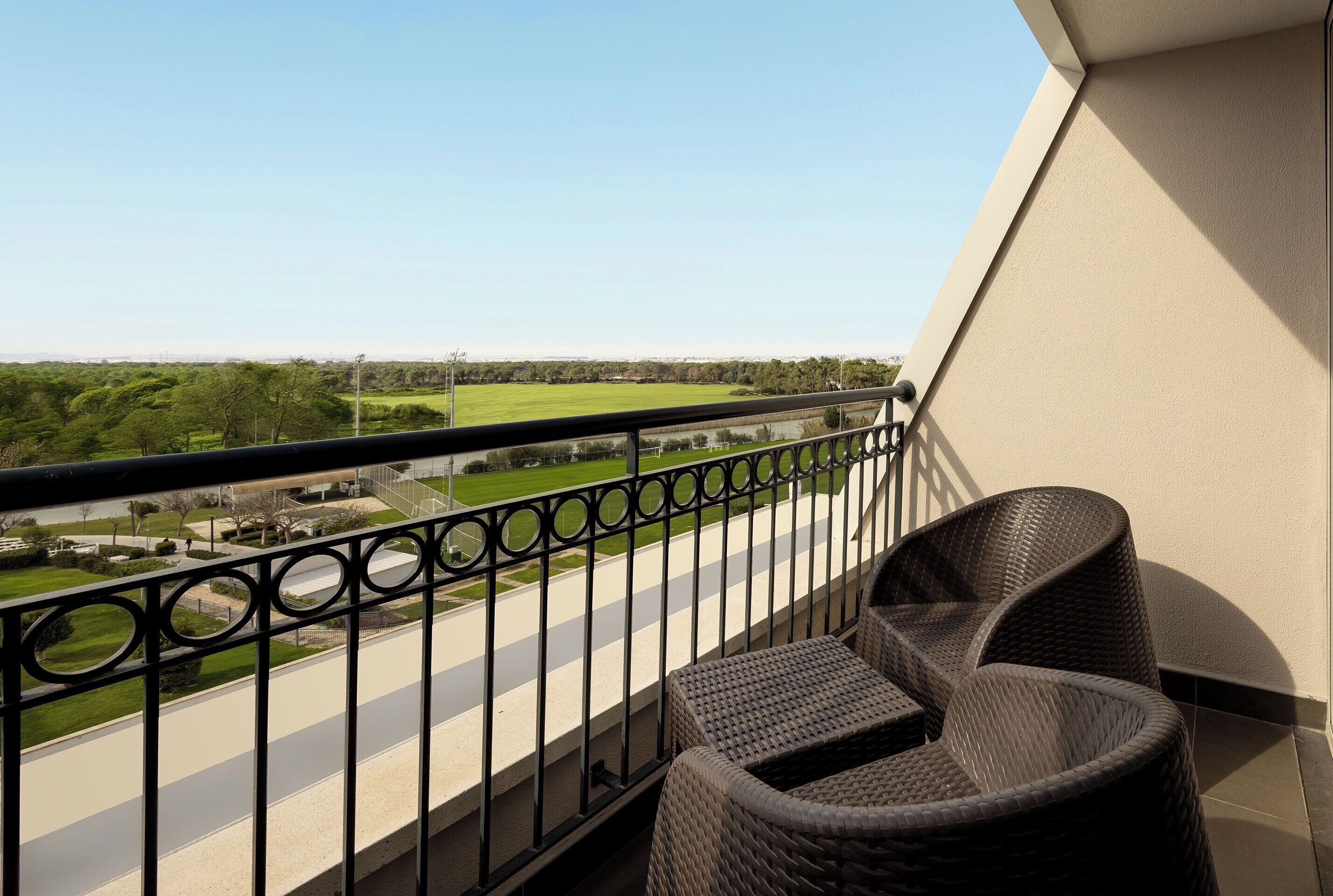 A balcony with wicker furniture, a metal railing, and a view of a green landscape and road.
