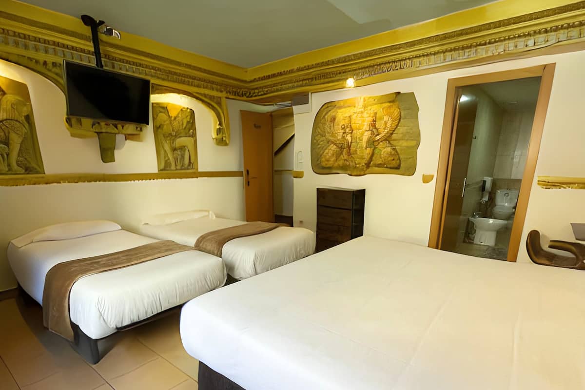 Habitación de hotel con dos camas individuales y una cama doble, un televisor montado en la pared y un baño visible a través de una puerta de cristal.