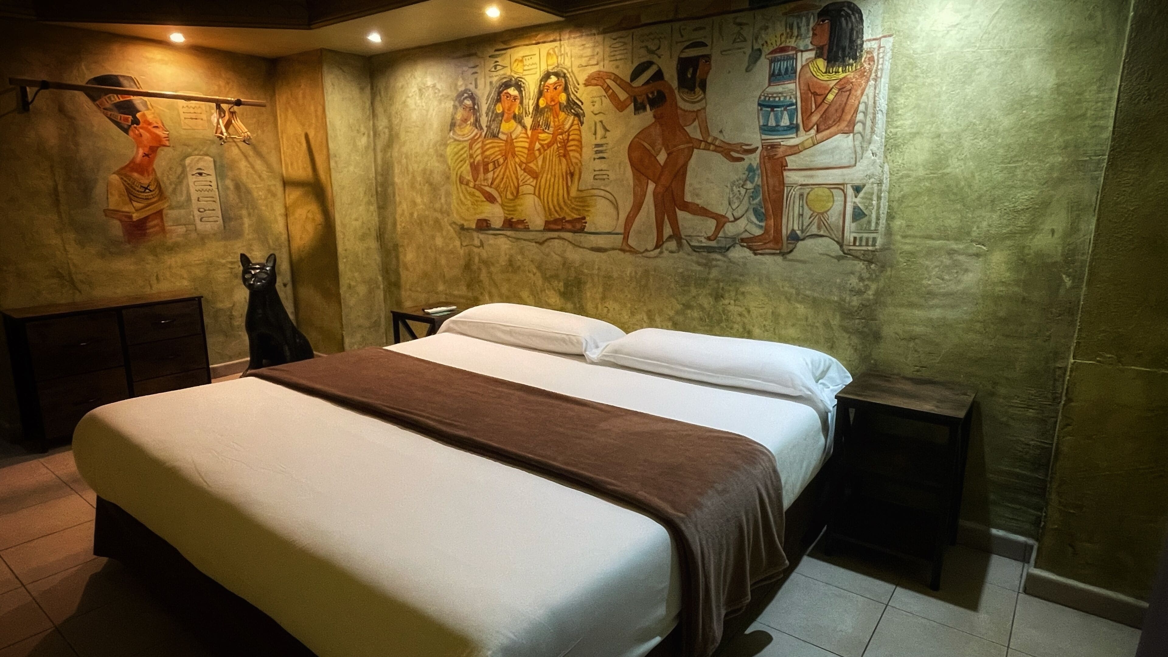 Une chambre d’hôtel avec un grand lit, un chat, une fresque murale et un petit bureau.