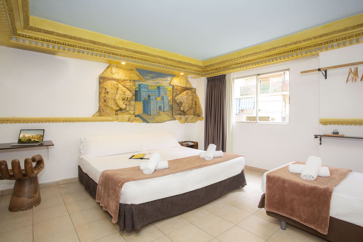 Una habitación de hotel con una cama grande, una cama más pequeña, un escritorio con una computadora portátil, un adorno en la pared y una ventana con cortinas.