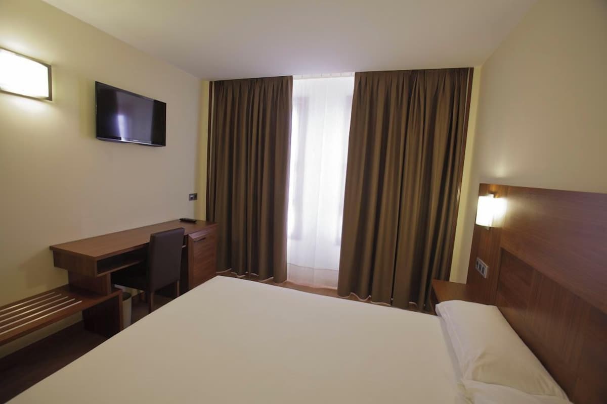 Habitació d'hotel amb llit, escriptori, cadira, televisió i cortines.
