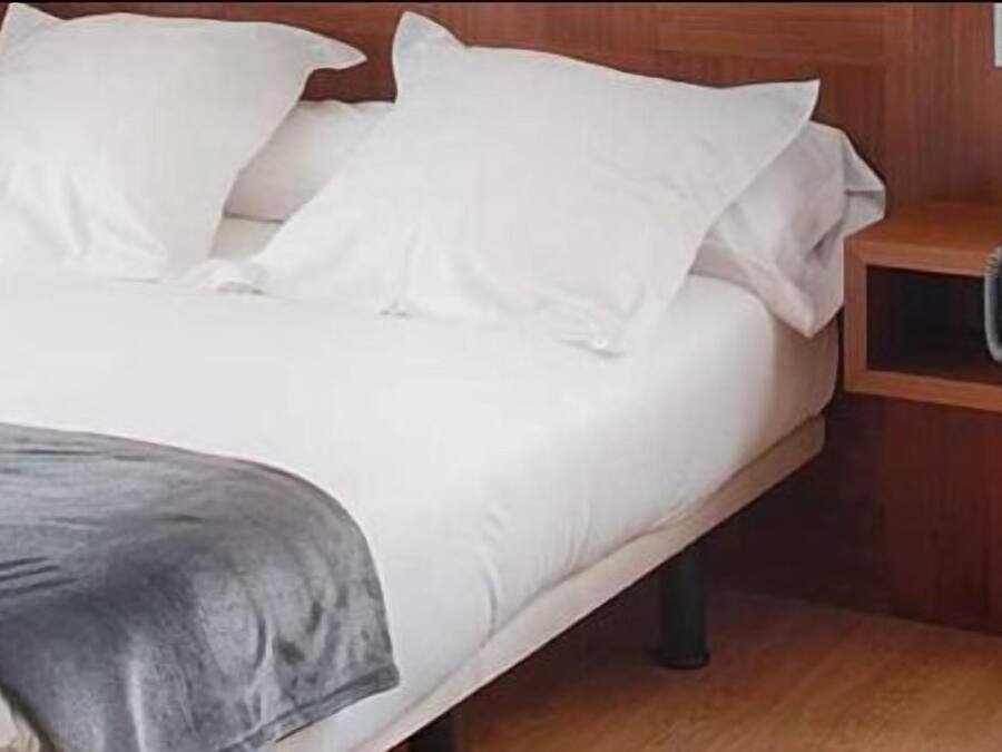 A neatly made bed with white linens and a gray throw blanket, a wooden nightstand with a telephone, and a wooden headboard.