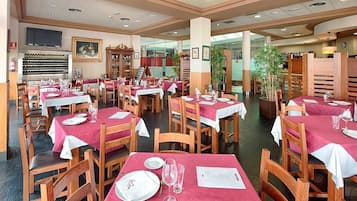 Un restaurante con varias mesas dispuestas, cubiertas con manteles blancos y servilletas rojas, sillas de madera y un televisor colgado en la pared.