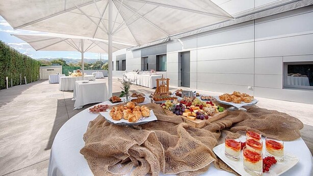 Un espace de restauration en plein air, avec des tables disposées sous de grands parasols, offrant une vue sur un bâtiment moderne et un jardin aménagé.