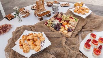 Une table sur laquelle sont disposés divers aliments, notamment des pâtisseries, des fromages et des fruits, le tout présenté sur une nappe blanche avec une toile de jute en dessous.