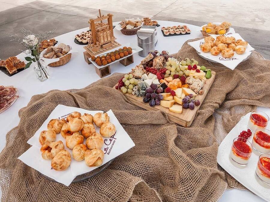 Una mesa con diversos alimentos como pasteles, quesos y frutas, dispuesta sobre un mantel blanco con un forro de arpillera.