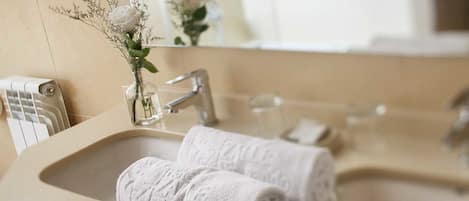 A bathroom with a beige sink, two rolled towels, a glass vase with a plant, and a mirror reflecting a window.