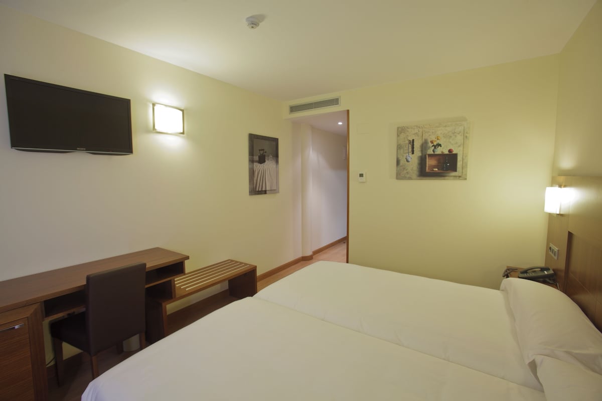 Una habitació d'hotel amb un llit, una taula de treball, una cadira, una televisió i una obra d'art petita a la paret.