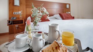 Un plateau de petit-déjeuner avec des tasses à café, une grande tasse, un verre de jus d’orange et une tranche de pain grillé, posé sur une table dans une chambre d’hôtel dotée d’une tête de lit en bois et d’un lit en arrière-plan.