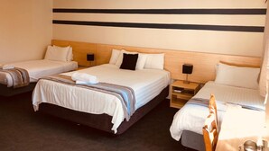 A hotel room with three single beds, a wooden headboard, bedside tables with lamps, and a ceiling light.