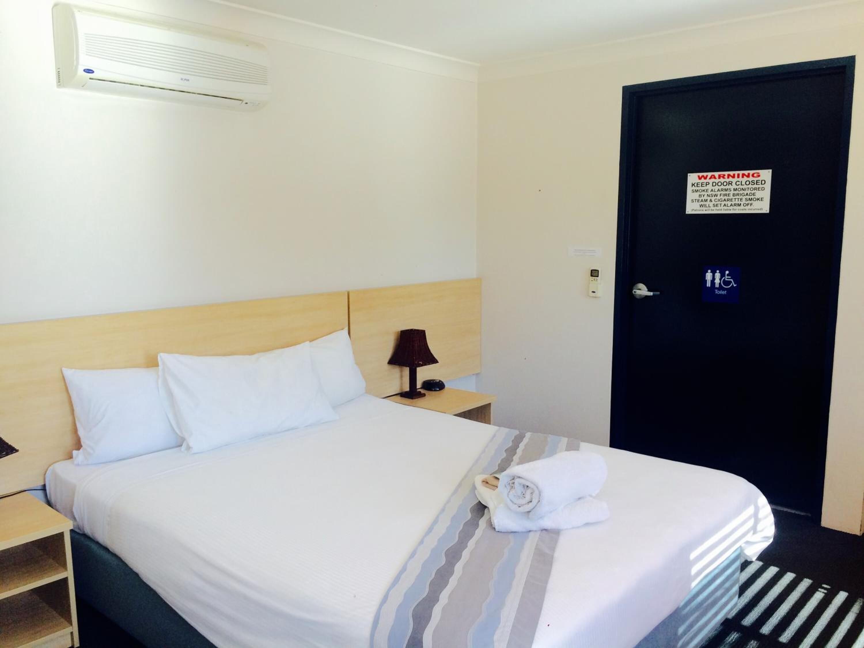 Habitación de hotel con cama, mesita de noche, lámpara, toalla, puerta con un letrero de advertencia y símbolo de inodoro.