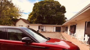 Ein rotes Auto vor einem Motel mit überdachter Außenbestuhlung und einem großen Baum im Hintergrund.