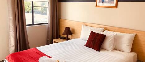 Une chambre d’hôtel avec un grand lit, une table de chevet, une lampe, un tableau encadré au mur et une fenêtre avec des rideaux.