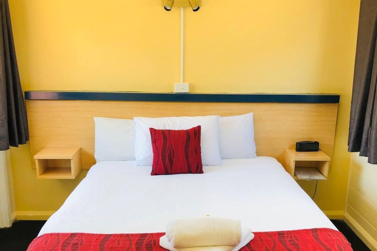 Ett hotellrum med en stor säng, två sängbord, ett litet bord, ett rött och svart mönstrat överkast, vita kuddar och en röd prydnadskudde.