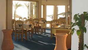 A dining area with wooden chairs and tables, large windows, and a view of palm trees. - Red Sea Relax (Dahab)