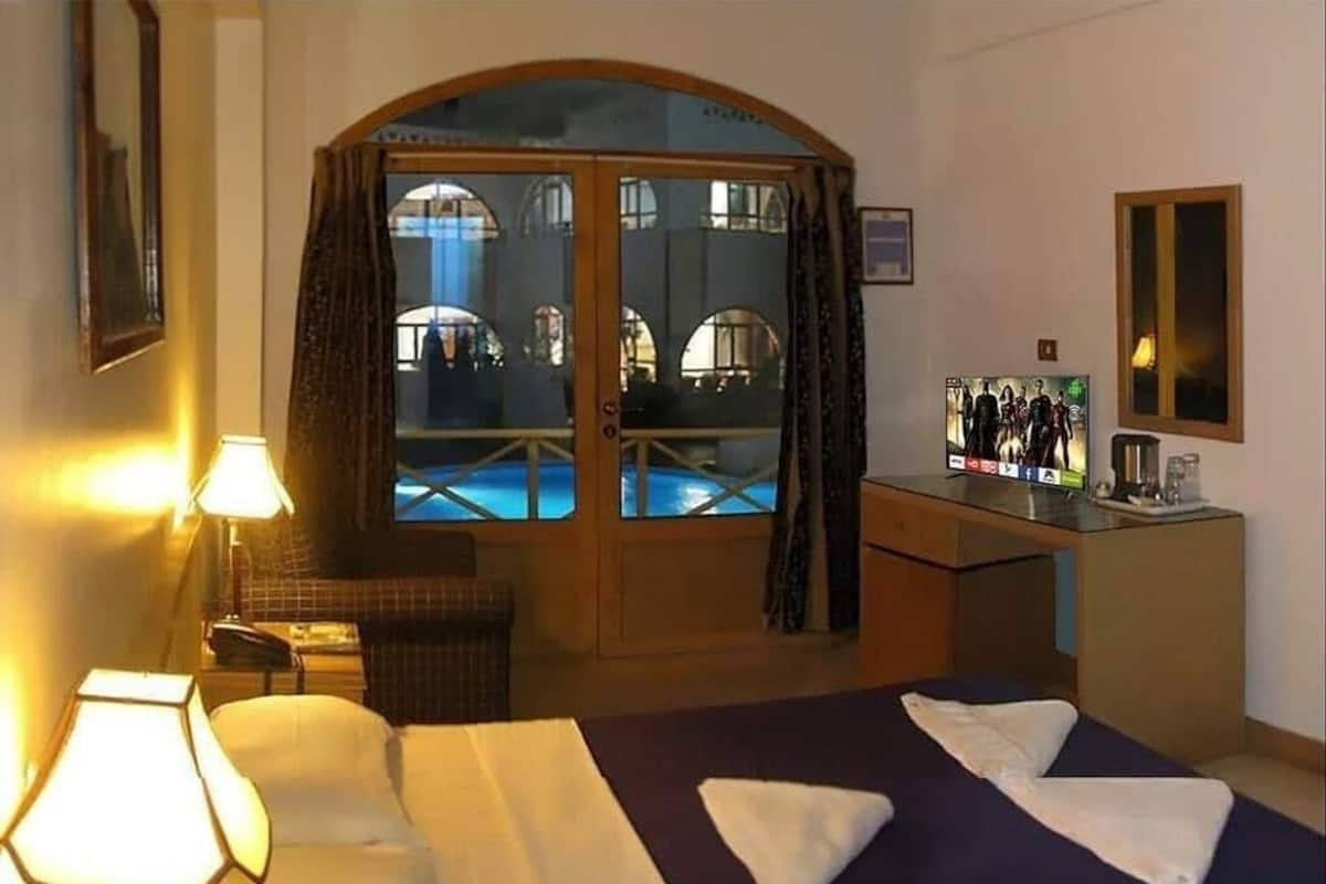 A hotel room with a bed, a desk, a chair, a lamp, and a view of an outdoor swimming pool through a glass door.