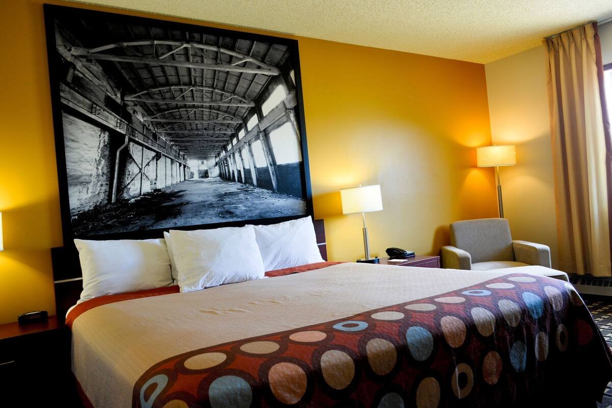 Habitación de hotel con una cama grande, dos lámparas de noche, una silla, un escritorio con teléfono y un mural grande en la pared.