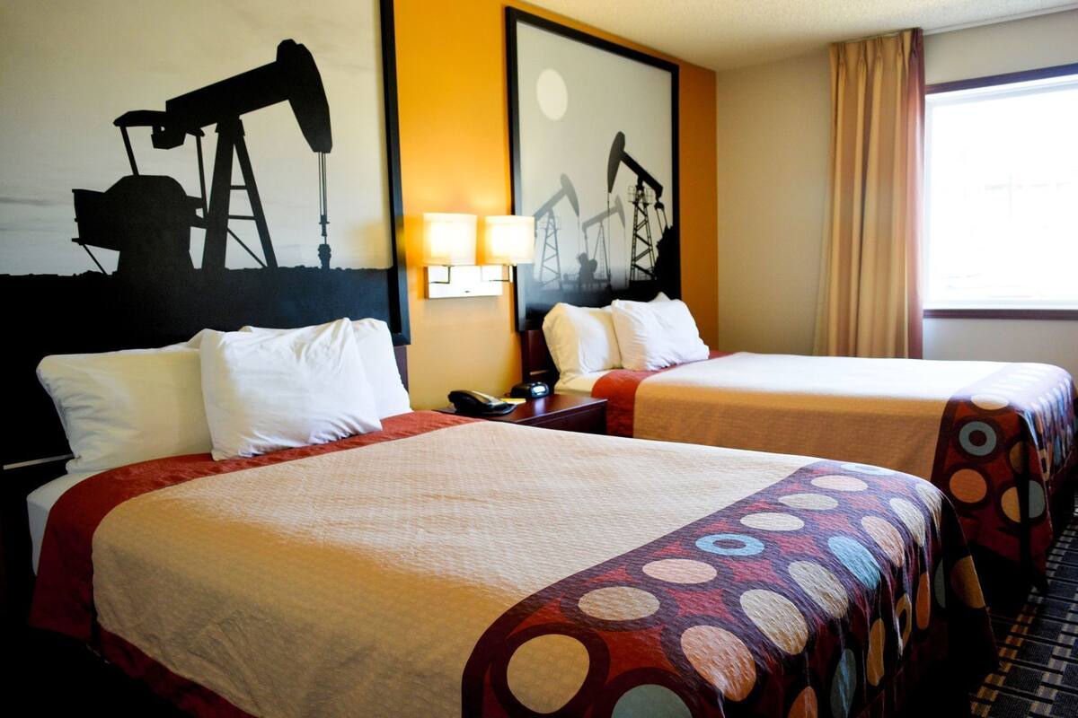 Habitación de hotel con dos camas, una mesita de noche, una lámpara y una obra de arte que representa bombas de petróleo.