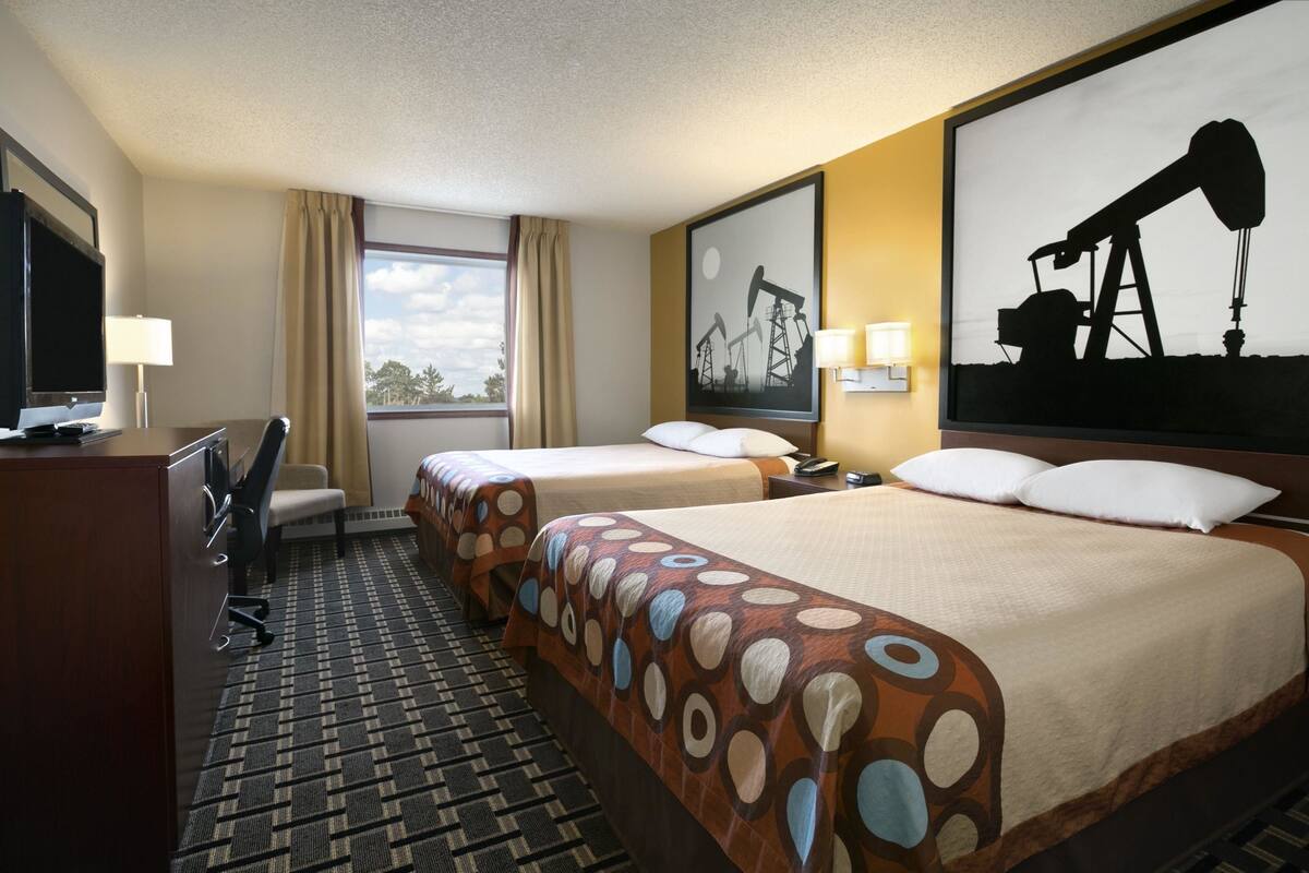 Habitación de hotel con dos camas, un escritorio con televisor, una silla, un ventanal con cortinas y un cuadro de torre de perforación de petróleo.