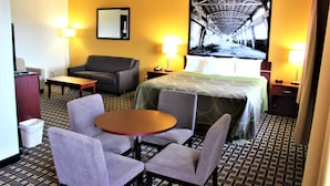 Ein Hotelzimmer mit einem Bett, zwei Sesseln, einem kleinen Tisch, einem Schreibtisch, einem Spiegel und einem an der Wand befestigten Fernseher.