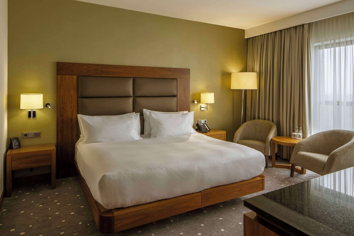 A hotel room with a large bed, two bedside tables with lamps, a small table with two chairs, and a large window with curtains.