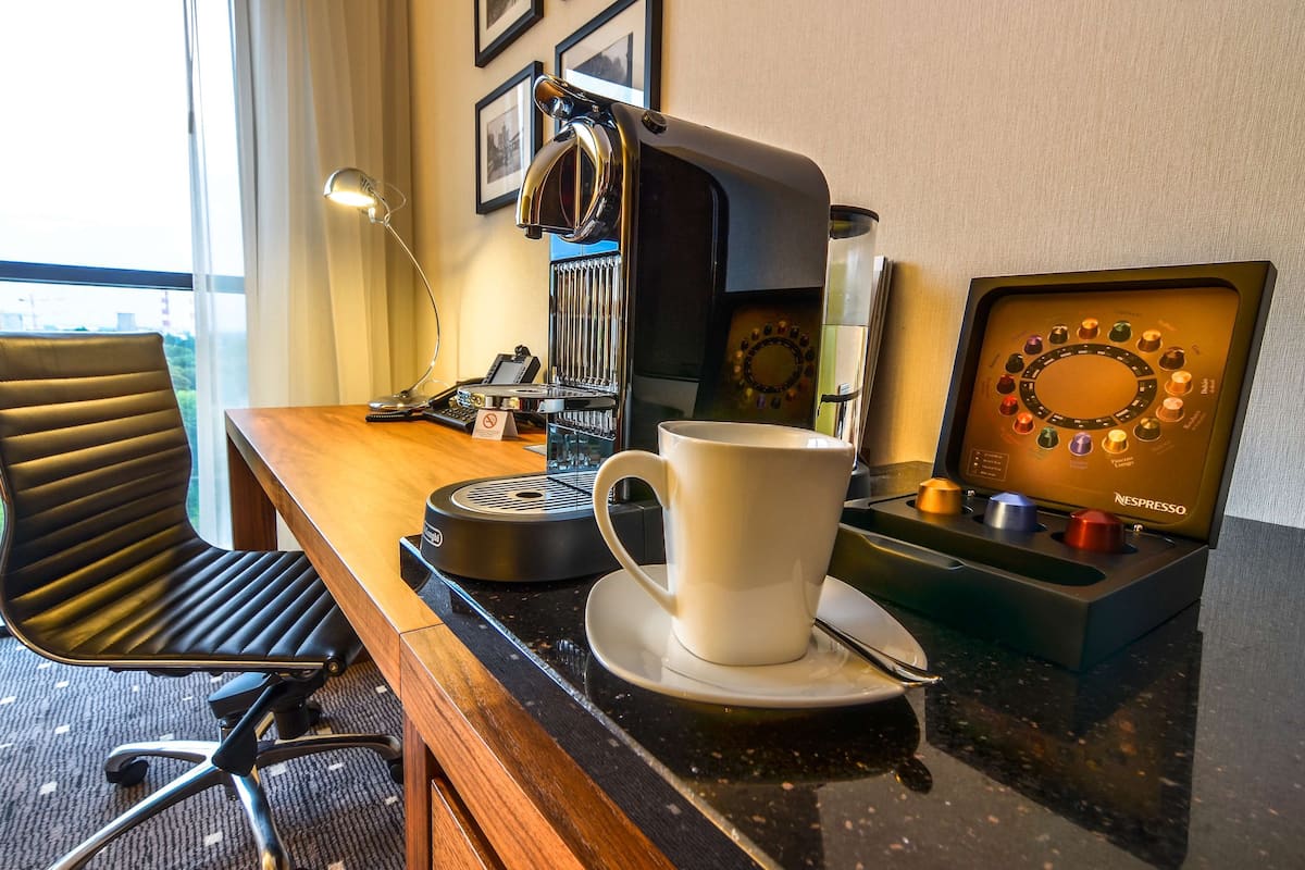 A hotel room with a wooden desk, a black office chair, a coffee machine, a cup of coffee, and a Nespresso coffee pod holder.