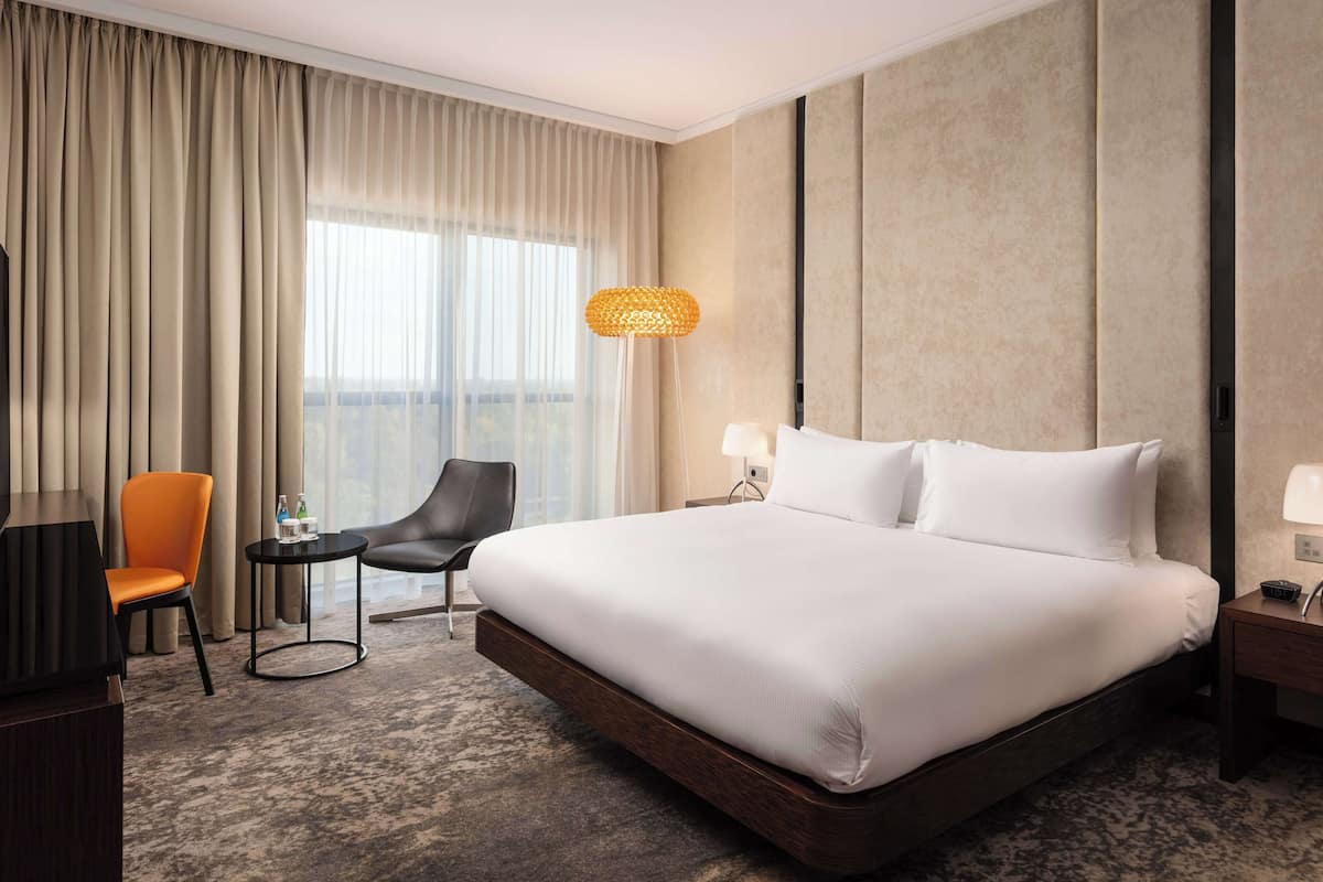 A hotel room with a large bed, a desk with a lamp, a chair, and a view of the city through the window.