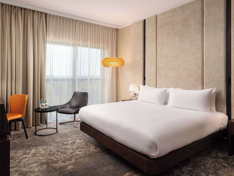 Habitación de hotel con una cama grande, un escritorio con lámpara, una silla y vistas a la ciudad desde la ventana.