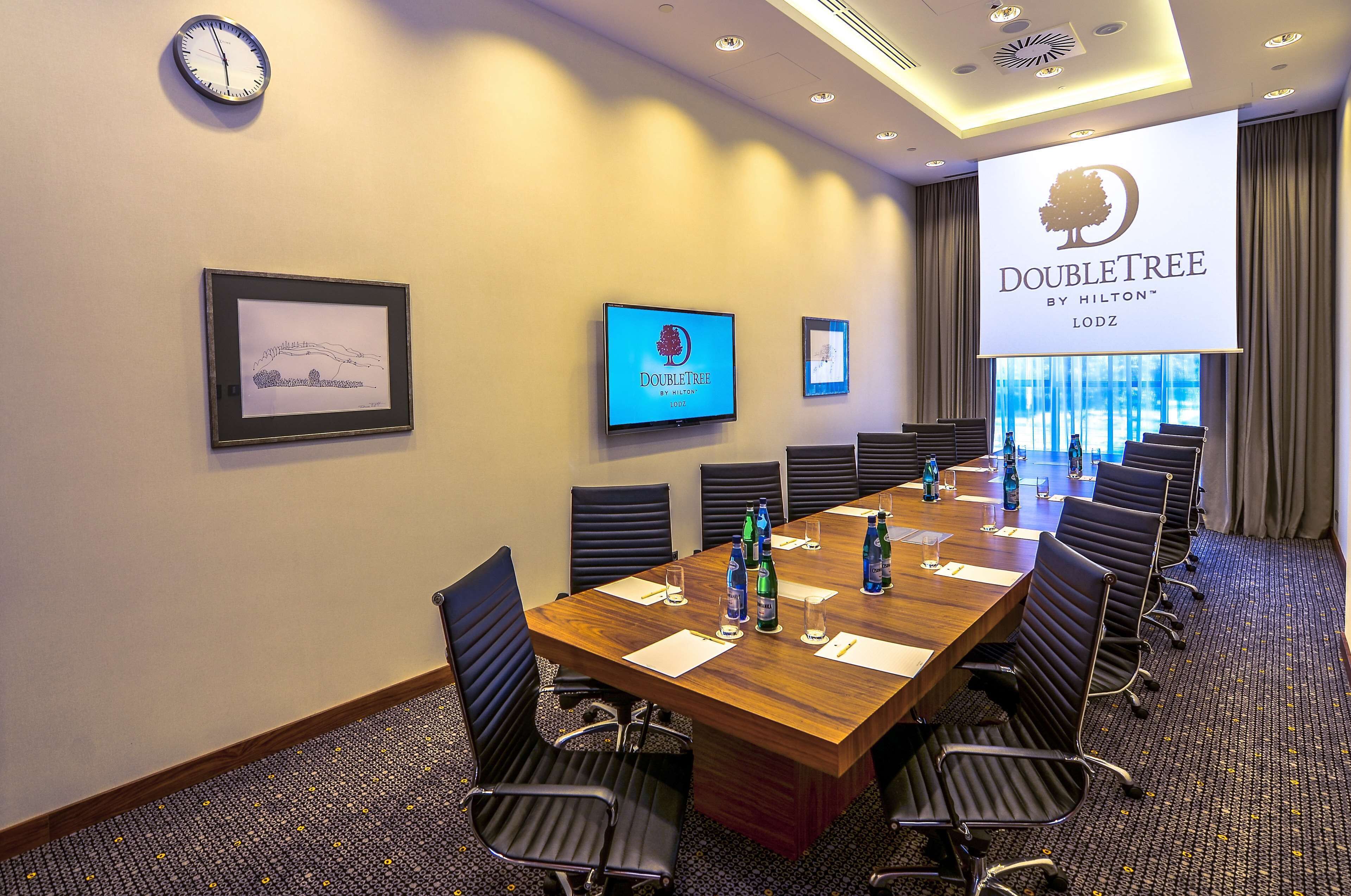 Un salón de conferencias con una larga mesa, sillas, un reloj, una pantalla de televisión y una pantalla de proyección que muestra el logotipo de DoubleTree.