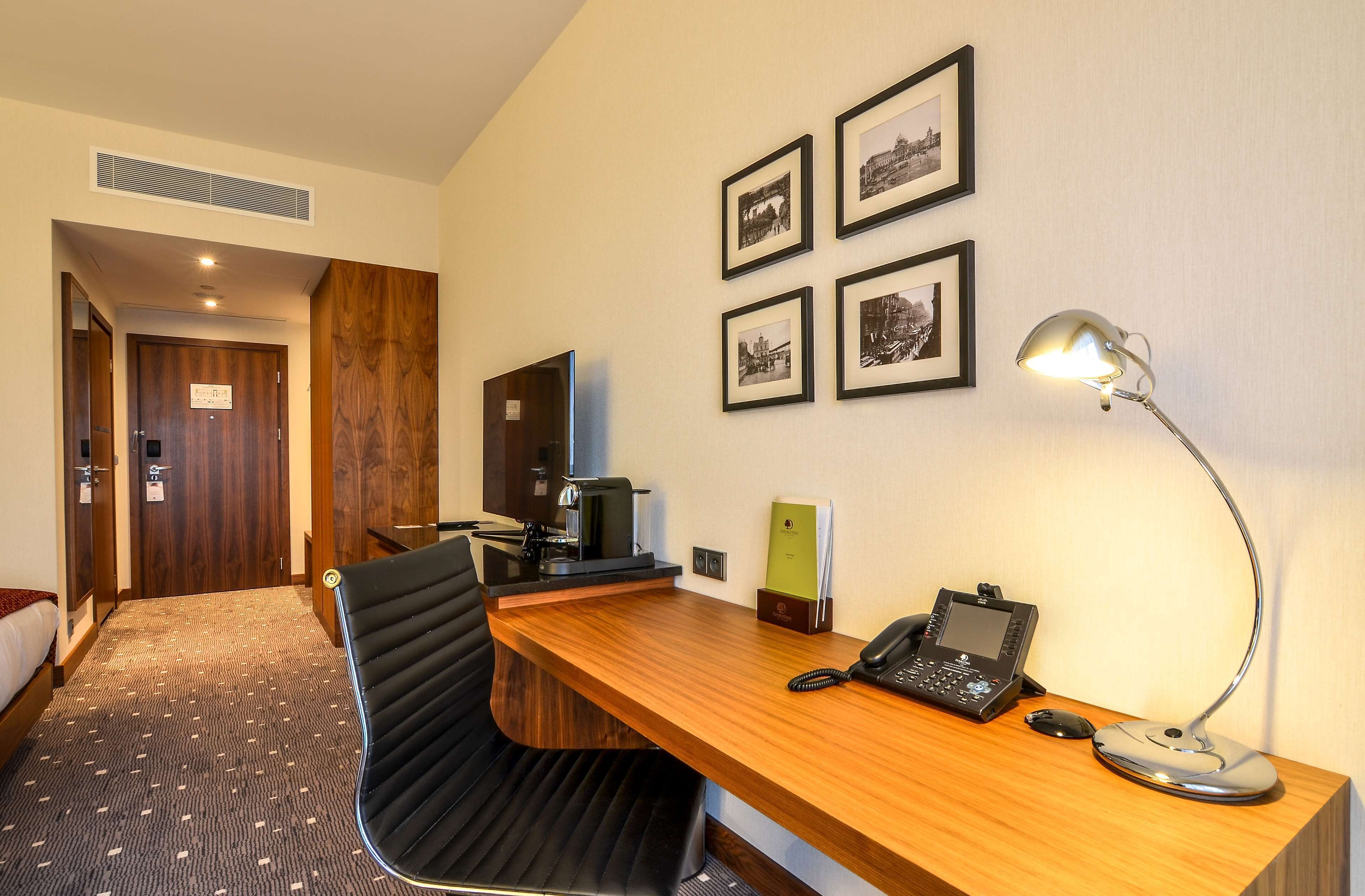 A hotel room with a wooden desk, a black office chair, a desk lamp, a telephone, and framed pictures on the wall.