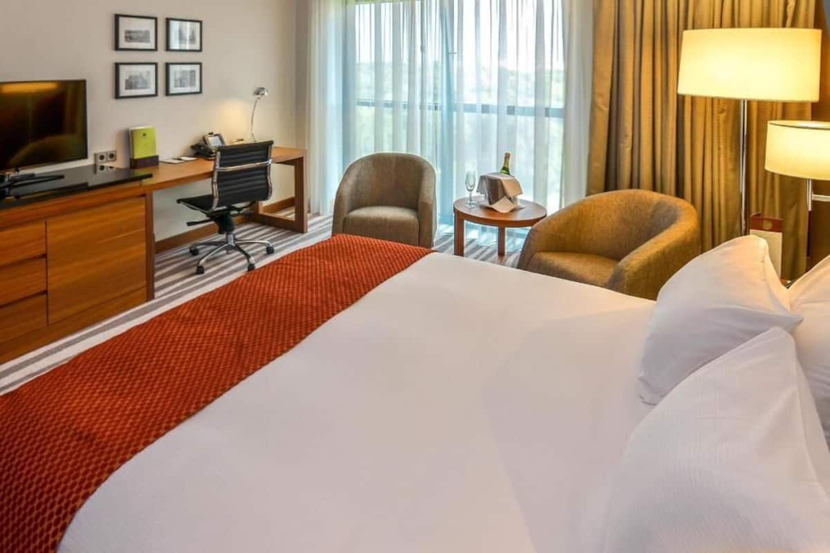 A hotel room with a large bed, a desk with a computer, two armchairs, a small table with a bottle, and a TV on a dresser.