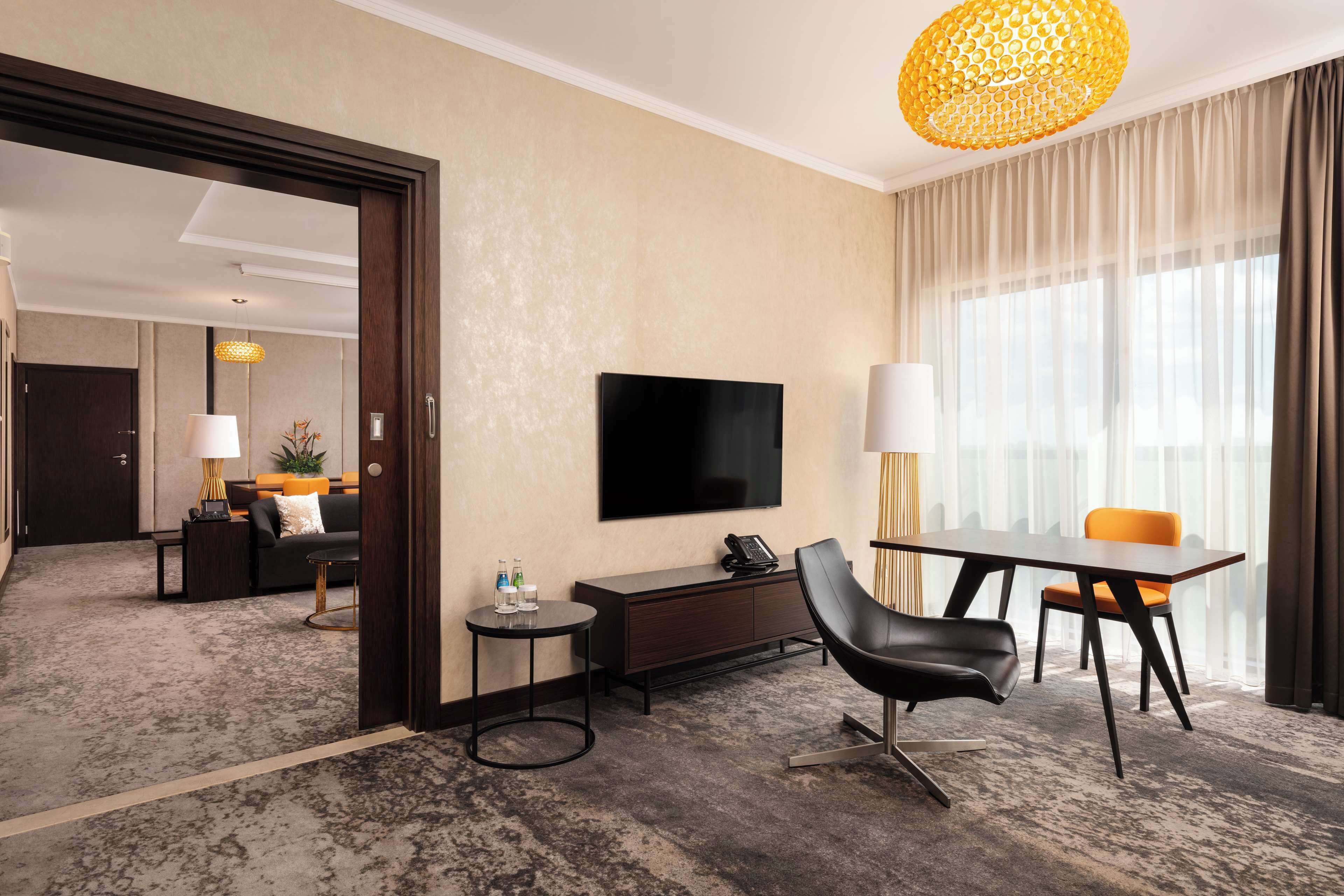 A hotel room with a flat-screen TV, a desk with a lamp, a dining area with a table and chairs, and a large window with curtains.