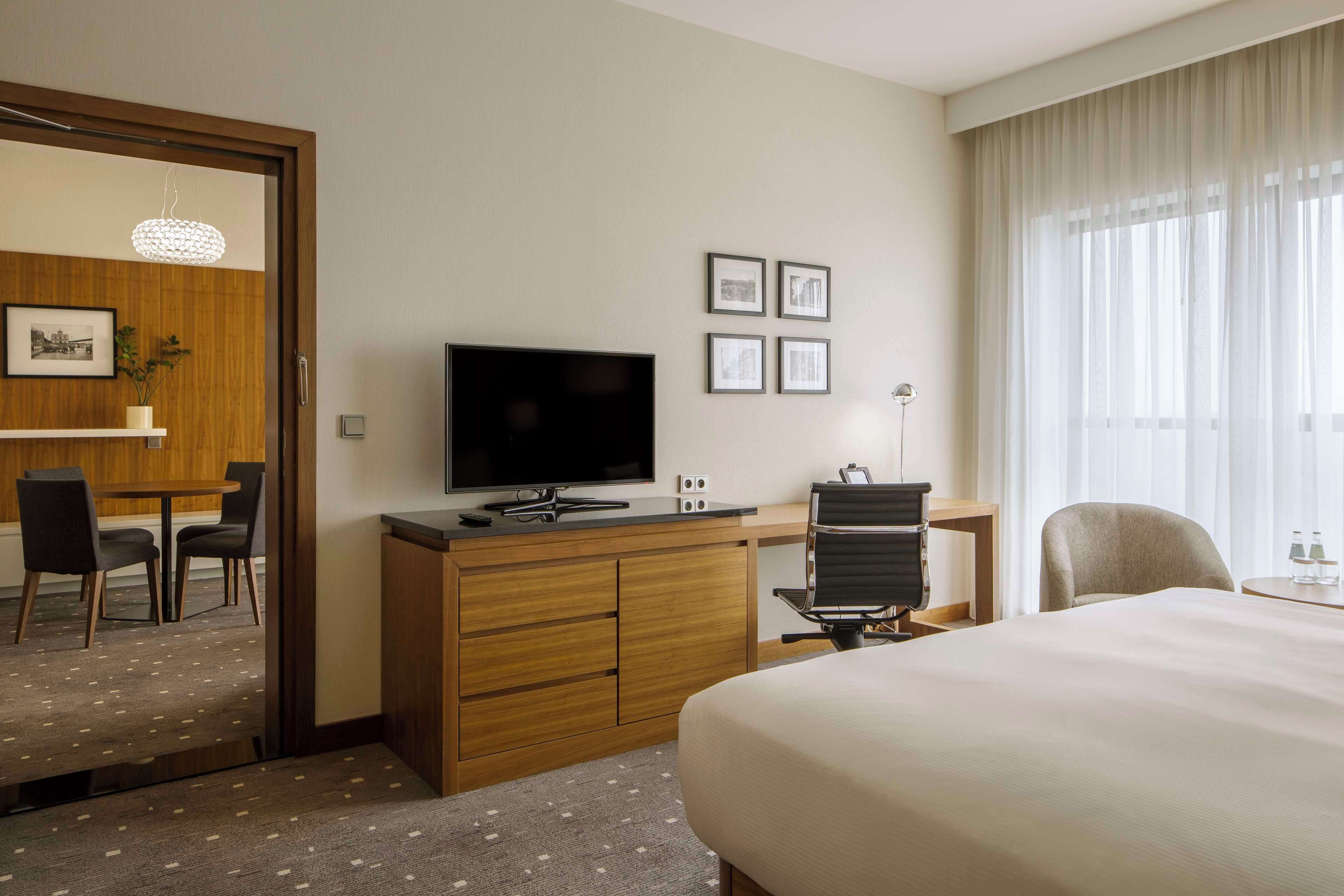 A hotel room with a bed, a desk with a computer, a TV, a chair, a small table with two glasses, and a window with curtains.
