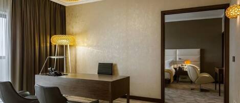 A hotel room with a wooden desk, two black chairs, a large floor lamp, and a bed with a white headboard.