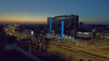 A large building with a prominent blue vertical feature, surrounded by a parking lot and a road with moving vehicles.