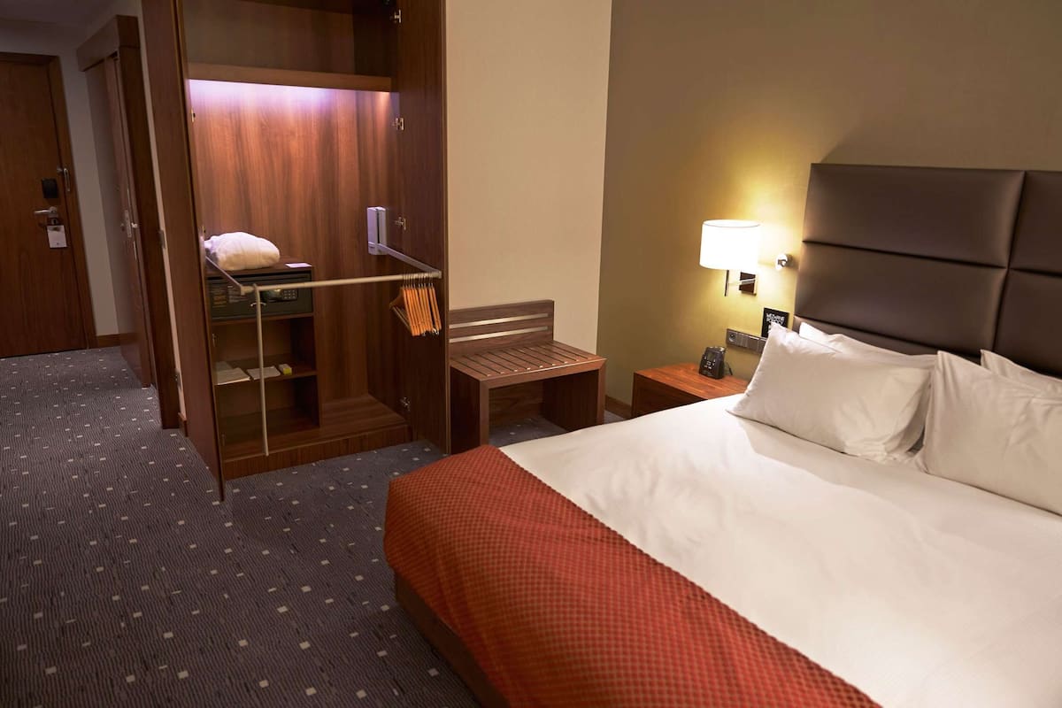 A hotel room with a bed, a wooden wardrobe, a bench, and a nightstand with a lamp.