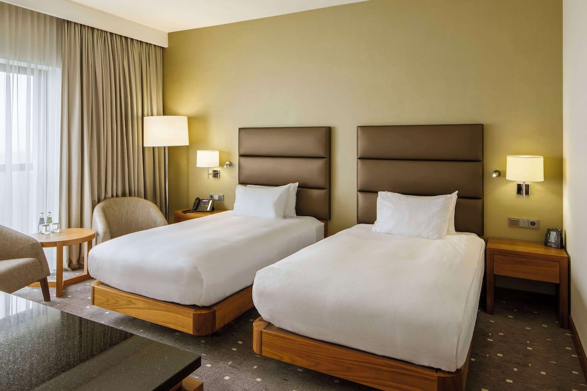 A hotel room with two beds, a desk, a chair, and a lamp.