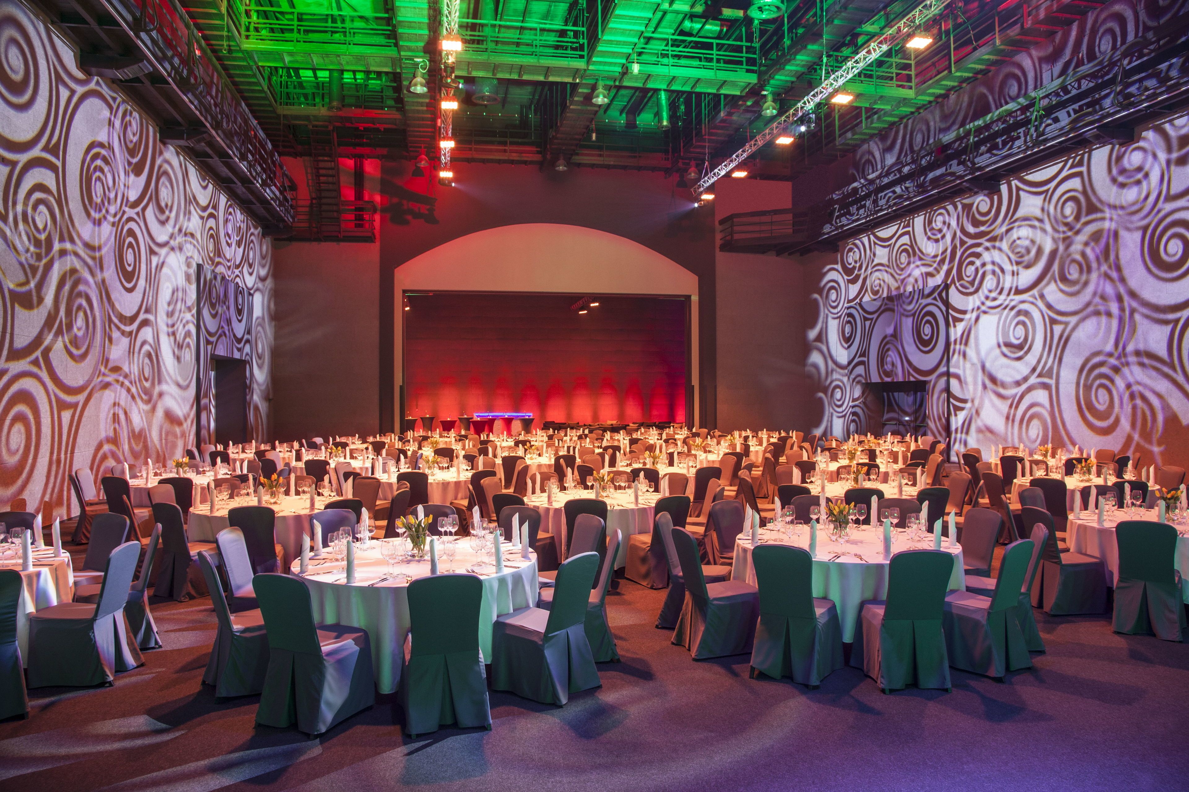 Une grande salle de banquet avec des tables rondes et des chaises, une scène et un décor mural à motifs.
