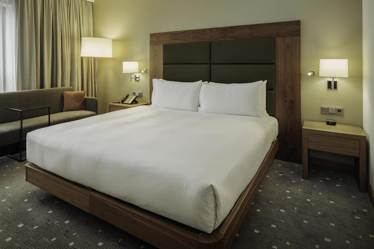 A hotel room with a large bed, a bedside table with a lamp, a nightstand, a sofa, and a window with curtains.