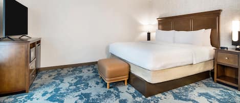 Habitación de hotel con cama, televisor, cómoda de madera, mesita de noche y alfombra azul estampada.
