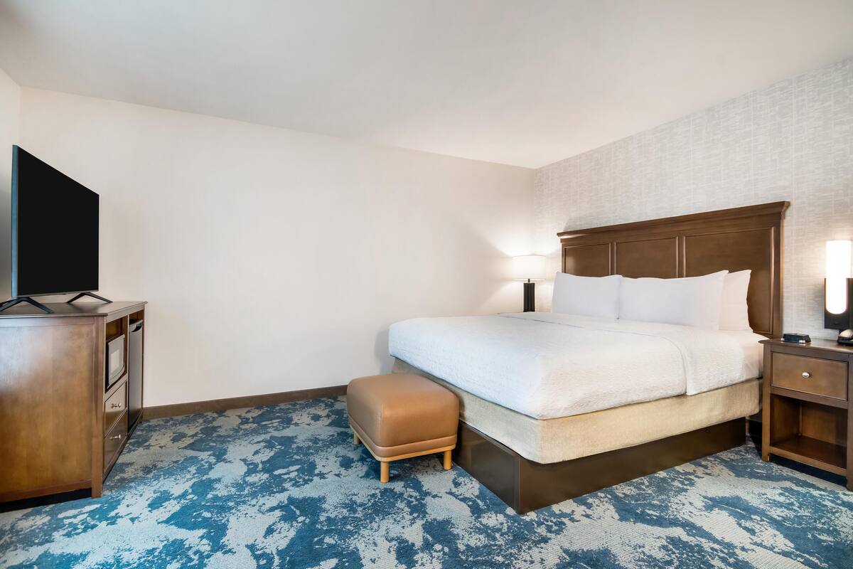 A hotel room with a bed, a TV, a wooden dresser, a nightstand, and a blue patterned carpet.