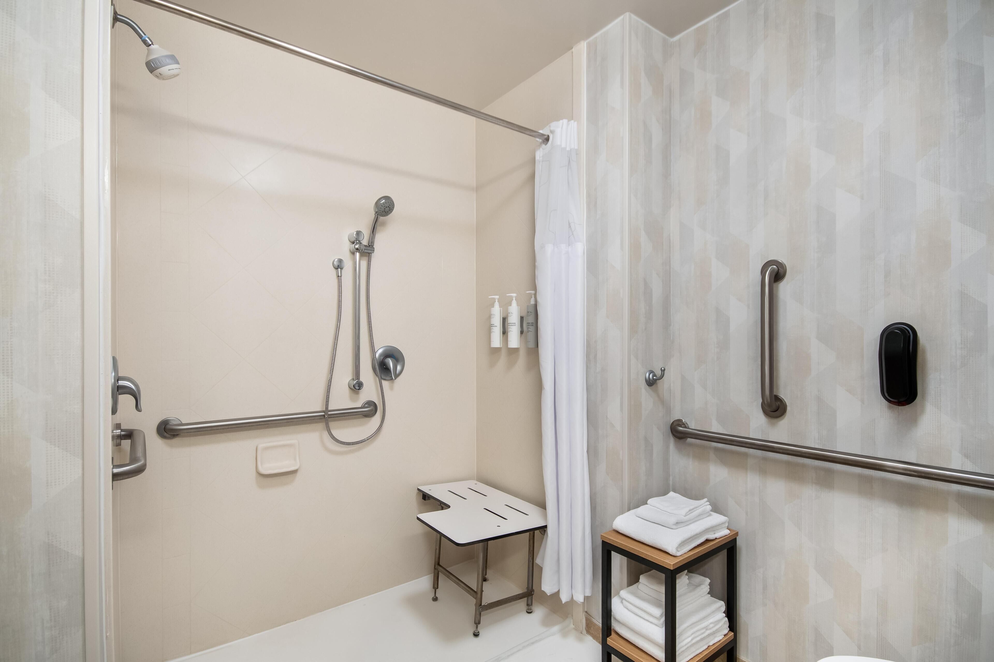 A modern bathroom with a shower, grab bars, a towel rack, and a small table.